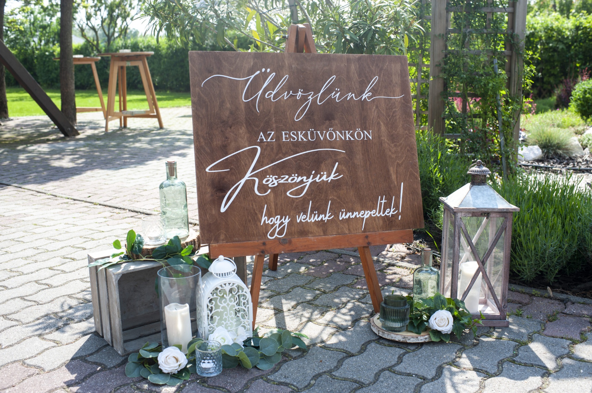 Rusztikus üdvözlő tábla lámpásokkal és gyertyákkal a kertben